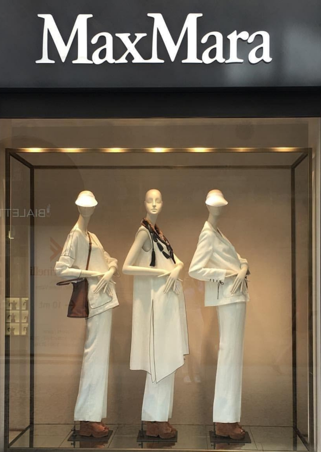 Βιτρίνα καταστήματος με τρεις γυναικείες κούκλες βιτρίνας σε λευκά σύνολα και minimal παρουσίαση μόδας.